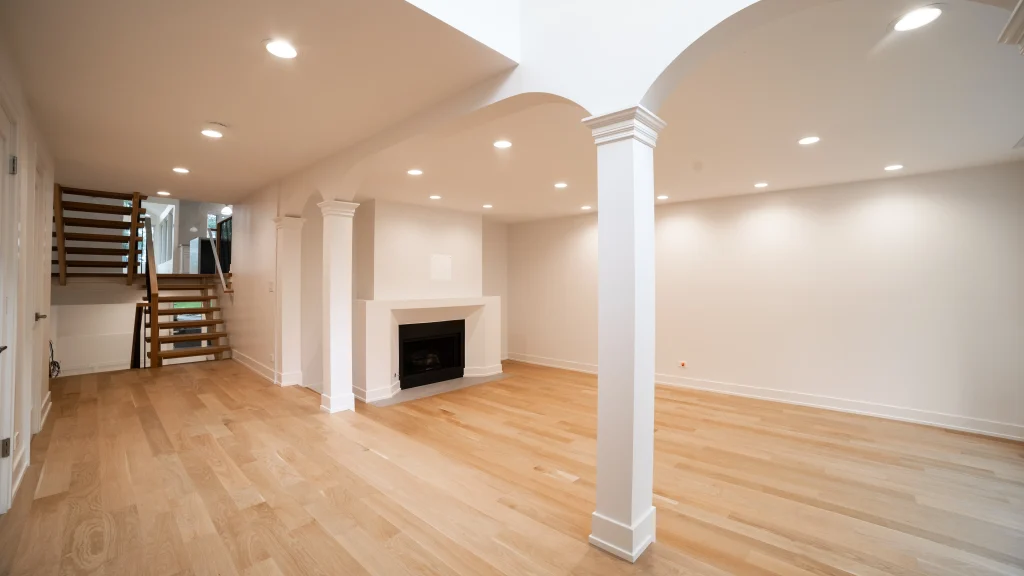 Spacious, empty basement with light wood flooring, white walls, recessed lighting, a white fireplace, and a wooden staircase leading upwards. Calm atmosphere