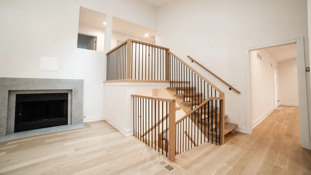 Modern living room with light wood flooring, a sleek gray fireplace, and open wooden staircase with black railings, conveying an airy, contemporary feel