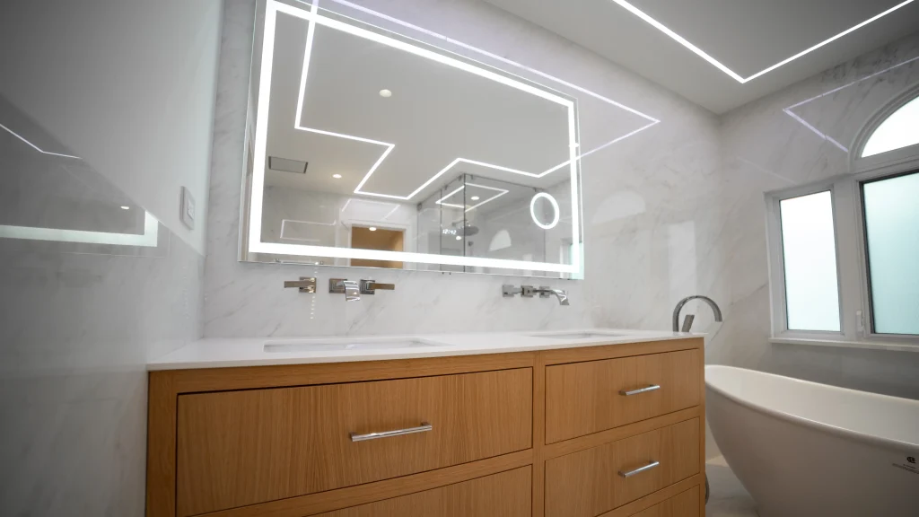 Modern bathroom with sleek white marble walls, featuring an illuminated rectangular mirror, a wooden double-sink vanity, and a white freestanding bathtub