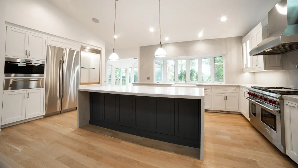 Spacious modern kitchen with hardwood flooring, white cabinets, large dark island, stainless steel appliances, pendant lights, and large windows