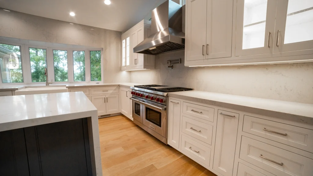 Spacious modern kitchen featuring stainless steel stove with red knobs, white cabinets, light wood floor, and large windows overlooking greenery