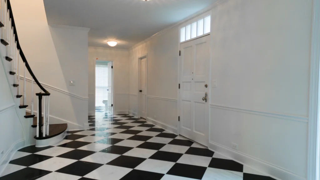 A spacious hallway with black and white checkered flooring, white walls, and a wooden staircase on the left