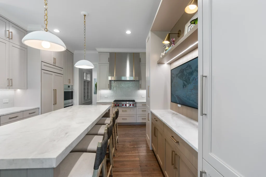 A modern kitchen features a long marble island with high chairs, pendant lights, sleek cabinetry, and a stainless steel stove, creating an elegant ambiance
