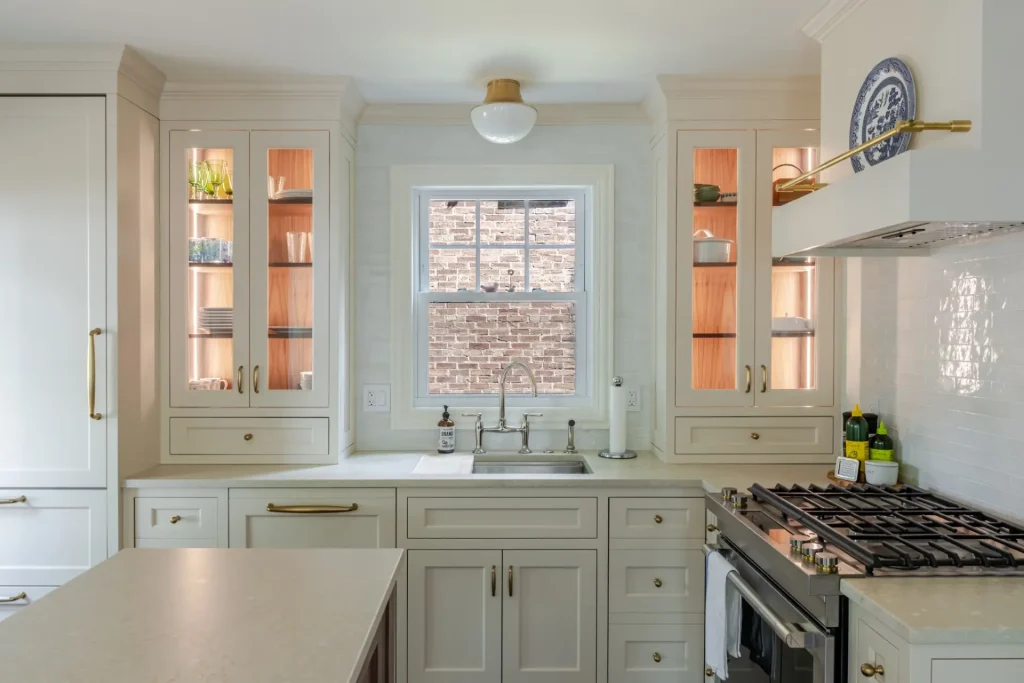 Bright kitchen with cream cabinets, glass-fronted displays, and a central window showcasing a brick wall