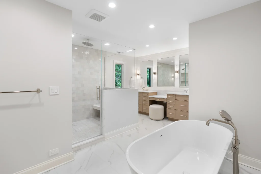 A modern bathroom with a freestanding white bathtub, glass shower enclosure, dual sinks on a wooden vanity, and bright overhead lighting