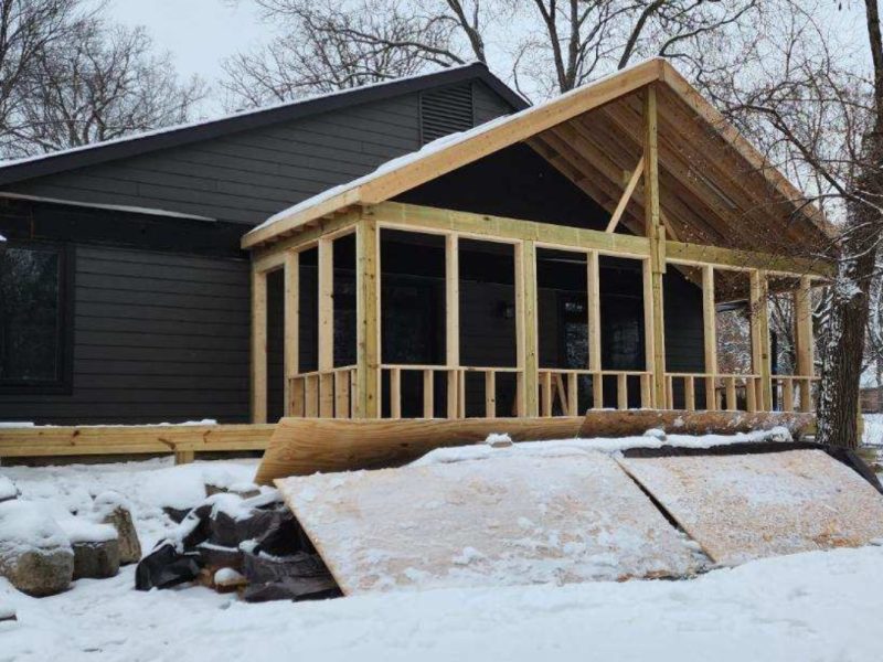 A dark gray house with a partially constructed wooden porch stands in a snowy landscape