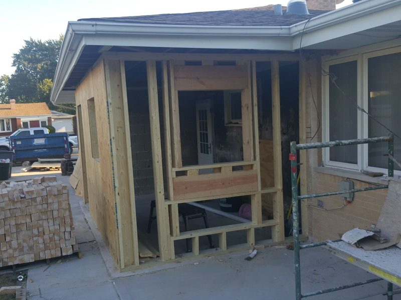 A house under renovation with an unfinished wooden porch frame, construction scaffolding, and a stack of bricks