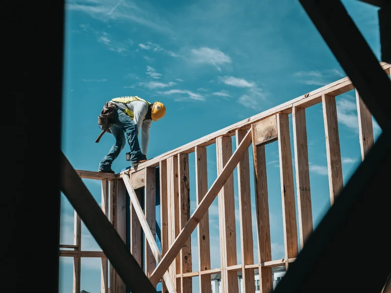 Construction worker wearing a helmet and safety vest, standing on a wooden frame against a blue sky