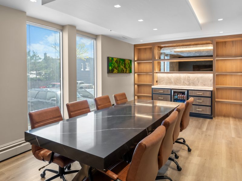 A modern conference room with a large, dark metallic table surrounded by eight brown leather chairs, wooden shelves, and bright natural light from large windows