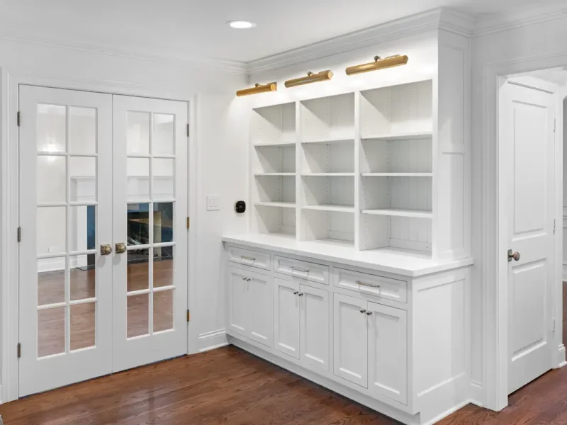 A bright, modern room with white built-in shelves and cabinets, wooden floor, and French doors