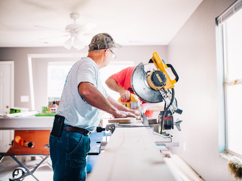 Two men work in a bright room, operating a circular saw on a long wooden plank