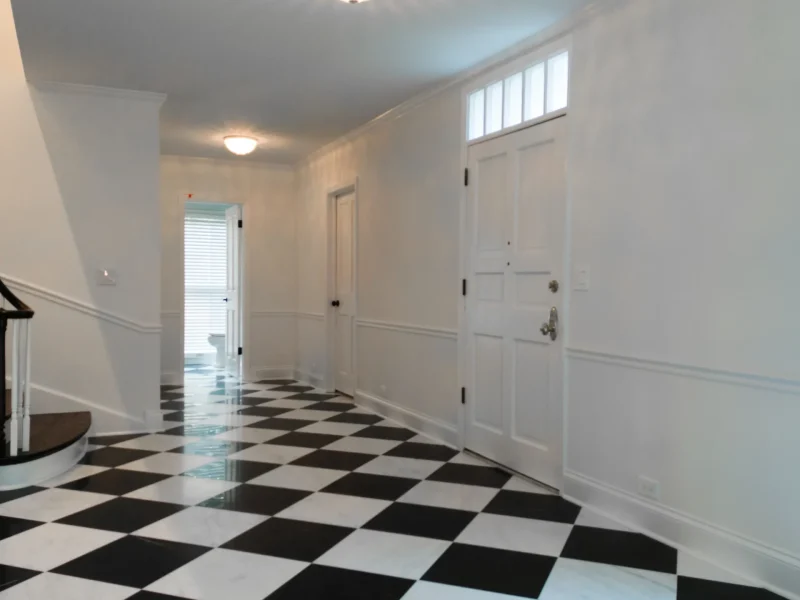 A spacious hallway with black and white checkered flooring, white walls, and a wooden staircase on the left